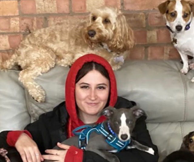 Beth, daycare manager, smiling on a sofa with a whippet in a harness, a cockapoo and a Jack Russell